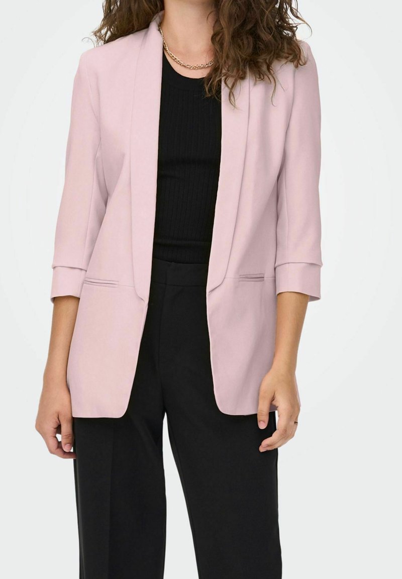 Blazer rosa claro con un diseño ajustado, dos bolsillos frontales y mangas remangadas, usado sobre una blusa negra de canalé y pantalones negros.