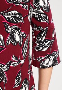 Tissu bordeaux avec un motif floral audacieux noir et blanc. Les manches courtes présentent un poignet uni et un détail de bouton pour la fermeture.