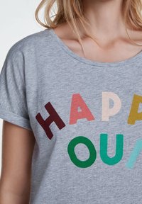 T-shirt gris clair à manches courtes avec une typographie multicolore écrivant "HAPPY" et "OUA" en gras, dans des polices variées. Texture de tissu douce.
