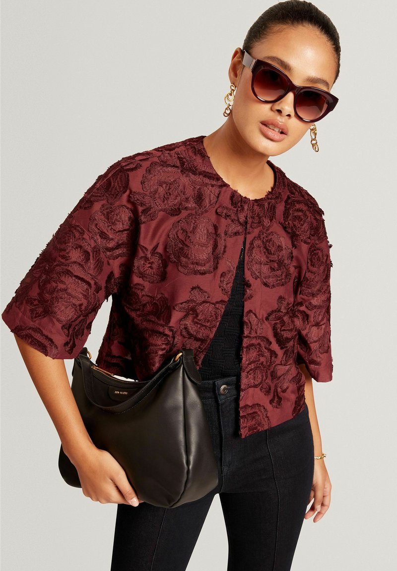 Veste bordeaux à texture florale, manches courtes et col rond ; associée à un sac à main noir. Le mannequin porte des lunettes de soleil surdimensionnées.