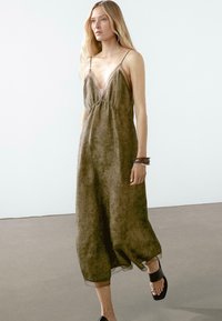 Olive green, sleeveless dress with a V-neck and elastic waist. Sheer overlay at the hem, lightweight fabric, and subtle texture. Flat black sandals.