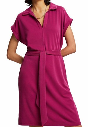 Femme portant une robe magenta courte, à manches courtes, longueur genou, avec un col et une ceinture en tissu nouée à la taille.