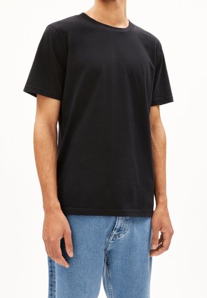 Mężczyzna w czarnej, gładkiej koszulce z krótkim rękawem i jasnoniebieskich dżinsach stoi na białym tle.