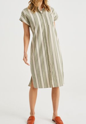 Žena v béžových šatech se zelenými vertikálními pruhy, krátkými rukávy, límečkem, přední kapsou a bočními rozparky, doplněná hnědými nazouvacími botami.