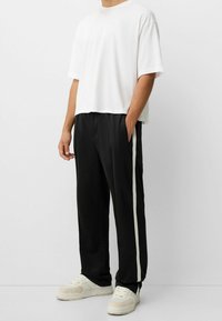 Bílý bavlněný tričko s uvolněným střihem, doplněné černými sportovními kalhotami s bílým bočním pruhem. Modelka má na sobě béžové tenisky.