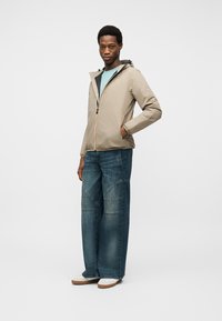 Νεαρός άνδρας που στέκεται, φοράει μια μπεζ ζακέτα με κουκούλα, ανοιχτό μπλε πουκάμισο, τζιν παντελόνι φαρδιάς γραμμής και λευκά αθλητικά παπούτσια σε λευκό φόντο.