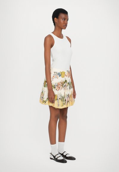 White fitted tank top with a pleated skirt featuring a floral print in pastel colors and a yellow hem. Black Mary Jane shoes and white socks.