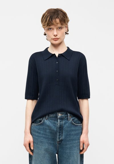 Jeune personne aux cheveux courts portant un polo à manches courtes côtelé bleu marine foncé et un jean en denim bleu, debout devant un fond blanc.