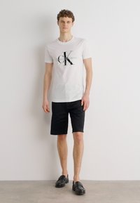 Balta kokvilnas T-kreklu ar melnu "Calvin Klein Jeans" logo, apvienota ar melniem īsiem biksēm un melniem mokasīniem, stāvot pret gaismas fona.