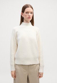 Sweter w kolorze miękkiej bieli, wykonany z dzianiny, z wysokim dekoltem, prążkowanymi detalami i długimi rękawami, w zestawieniu z beżowymi spodniami.