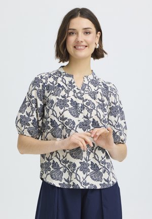 Mladá žena sa usmieva, má na sebe bielu blúzku s tmavými kvetinovými vzormi a nariasenými krátkymi rukávmi, spojenú s tmavomodrými nohavicami.