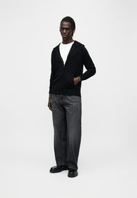 Sweat à capuche noir à fermeture éclair, avec capuche réglable, porté sur un t-shirt blanc, assorti à un jean gris foncé ample et des chaussures noires.