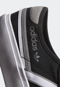 Musta kangaskenkä harmailla yksityiskohdilla, valkoisilla raidoilla ja tekstuurilla varustetulla pohjalla. Varustettu brodeeratulla brändäyksellä ja matalaprofiilisella muotoilulla.