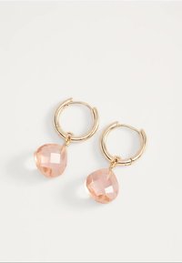 Kultaiset rinkkorengaskorvakorut, joissa on fasetoidut, läpikuultavat persikanväriset sydämenmuotoiset riipukset valkoisella taustalla.