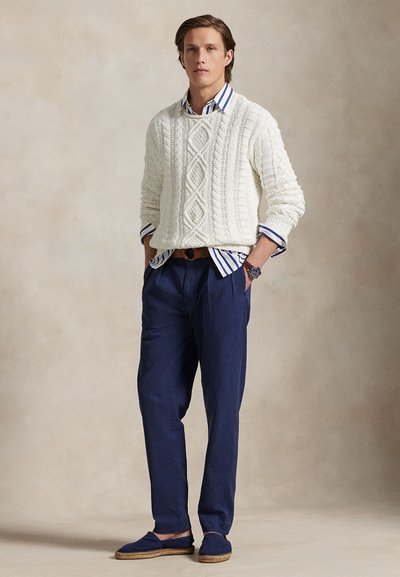 Mlad muškarac u bijelom džemperu s kabelskim uzorkom preko plavo-prugaste košulje, tamnoplavim hlačama, smeđim remenom i plavim espadrilama, stoji s rukama u džepovima.