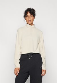 Pullover in pile beige con collo alto e zip anteriore a metà, abbinato a pantaloni della tuta scuri con vita regolabile.