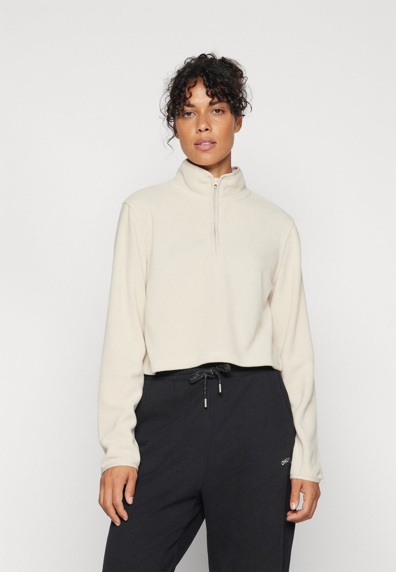 Pullover in pile beige con collo alto e zip anteriore a metà, abbinato a pantaloni della tuta scuri con vita regolabile.