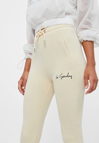 Světle béžové tepláky vyrobené z měkkého materiálu s pasem na šňůrku, obsahující boční kapsu a černý zakřivený text "You Someday" na noze.