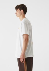 Bijela pamučna majica kratkih rukava s zlatnim tekstom sprijeda i straga, u paru s smešnim hlačama s vertikalnim prugama boja.