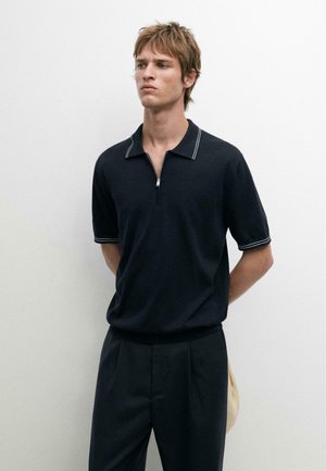 Młody mężczyzna ubrany w czarną koszulkę polo z krótkim rękawem i białym wykończeniem, czarne spodnie, stoi z rękami za plecami na tle jednolitej białej ściany.