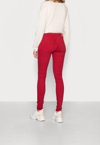 Mujer vistiendo jeans skinny rojos, suéter blanco de lana suave y zapatillas blancas gruesas, de pie contra un fondo gris claro liso.