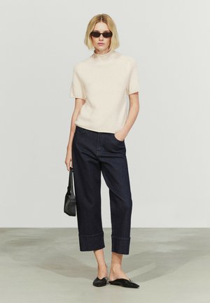 Frau trägt einen cremefarbenen Pullover mit kurzen Ärmeln, dunkle, weit geschnittene Jeans mit aufgekrempeltem Saum, schwarze Slipper, Sonnenbrille, hält eine kleine schwarze Handtasche, steht auf grauem Boden.