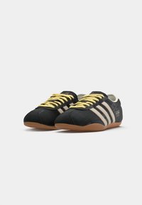 Black athletic shoes with yellow laces, featuring three beige stripes, a textured upper, and a gum rubber sole. Notable plaid pattern.