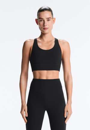 Athletische Frau mit zurückgekämmtem Haar, die einen schwarzen Sport-BH und hoch taillierte Leggings trägt und vor hellem einfarbigem Hintergrund steht.