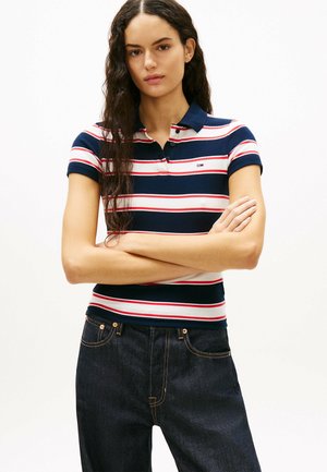 Kobieta o długich kręconych włosach stoi z założonymi na piersi rękami, ubrana w granatową, białą i czerwoną koszulkę polo w paski oraz ciemne dżinsy.