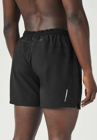 Vue de dos d'une personne portant un short de sport noir avec une ceinture élastique et une petite poche zippée à l'arrière, côté droit.