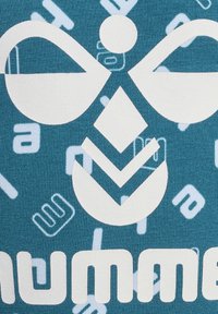 Teal tyg med vita geometriska och textmönster. Den centrala designen har ett stiliserat ansikte med cirkulära och triangulära former.