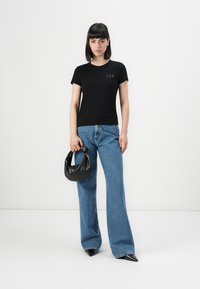 Una donna è in piedi contro un muro bianco, indossa una t-shirt nera della GAP, jeans a tutta gamba blu, scarpe nere con la punta e tiene in mano una borsa nera con un manico annodato.