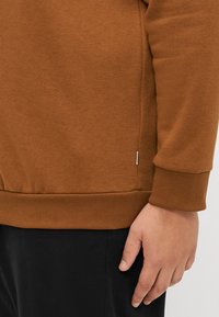 Bruine sweatshirt met een geribbelde zoom en manchetten, voorzien van een zijsplit detail. Materiaal heeft een zachte textuur. Hand zichtbaar aan de onderkant.