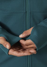 Chaqueta de color azulado con cremallera y un patrón texturizado y acolchado; agujeros para los pulgares en los puños. Presenta una construcción de hombros sin costuras y una cremallera completa.