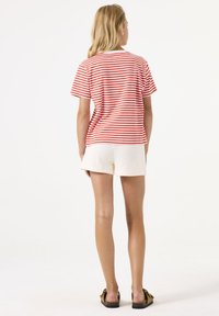 Camiseta de manga corta a rayas rojas y blancas, de corte relajado, combinada con pantalones cortos de textura crema y sandalias marrones. Fondo plano.