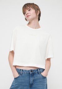 Valkoinen lyhythihainen crop-top -paita pehmeää kangasta, jossa on kevyesti värjätty "REPLAY"-brändin nimi brodattuna rinnassa.