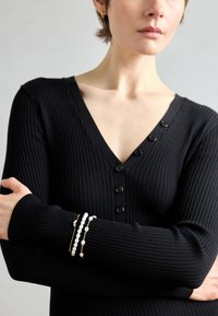 Top negro de manga larga con canalé, con escote en V y detalle de botones. Dos pulseras en capas, una con cuentas blancas y otra con acentos dorados.