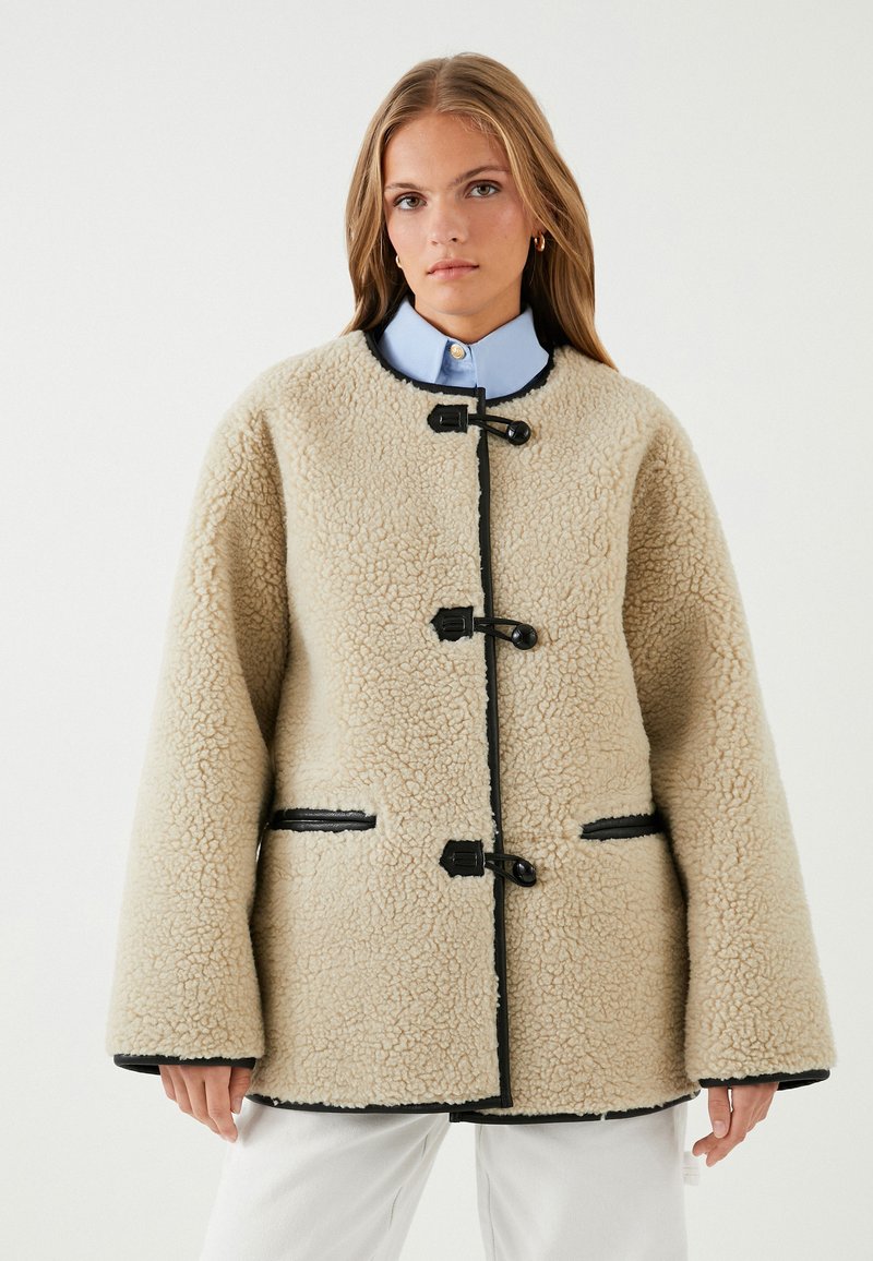 Chaqueta de forro polar beige con una superficie texturizada, ribetes negros, cierres tipo toggle y bolsillos laterales; cuenta con un collar redondeado y mangas amplias.
