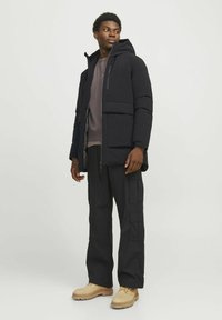 Sort isoleret jakke med hætte, to frontlommer, lagdelt over en grå sweatshirt. Parret med mørke cargo bukser og beige støvler.