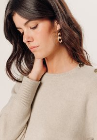 Beiger Strickpullover mit gerippten Bündchen und Rundhalsausschnitt, mit dekorativen Knöpfen an der Schulter und strukturiertem Stoff. Goldene Creolen.