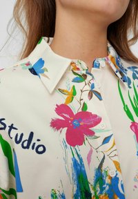 Bloes met bloemenprint in lichtcrème stof met levendige roze, blauwe en groene bloemen. Heeft een klassieke kraag en witte knopen.