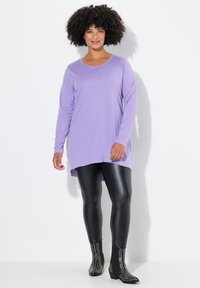 Langärmliges lila Oberteil mit rundem Ausschnitt, kombiniert mit glänzenden schwarzen Leggings und mit Nieten besetzten Stiefeletten, vor einem weißen Hintergrund.