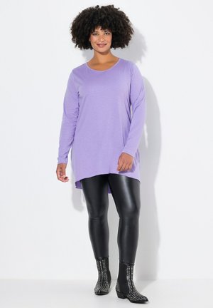 Langärmliges lila Oberteil mit rundem Ausschnitt, kombiniert mit glänzenden schwarzen Leggings und mit Nieten besetzten Stiefeletten, vor einem weißen Hintergrund.