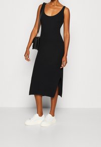Czarna prążkowana sukienka z okrągłym dekoltem i rozcięciem z boku, zestawiona z białymi trampkami. Sukienka jest dopasowana i sięga do połowy łydki.