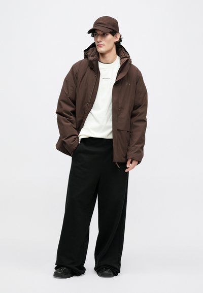 Brūna izolēta jaka ar kapuci, vilkta pār baltu T-kreklu, kopā ar vaļīgām melnām biksēm un atbilstošu brūnu cepuri. Melni apavi.