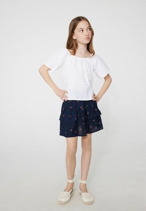 Niña joven con pelo largo que lleva una blusa blanca de manga corta, falda azul marino con volantes y estampado de cerezas, y zapatos blancos atados al tobillo.