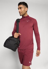Bordowy sportowy longsleeve z półzamkiem, w połączeniu z pasującymi szortami. Trzyma czarną torbę z paskiem na ramię. Białe akcenty logo.