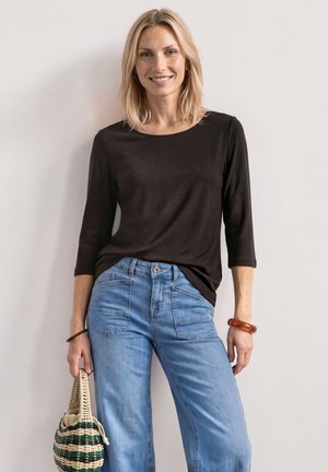 Blonde Frau mit schwarzem Langarmshirt und blauer Jeans, hält eine gestreifte gewebte Tasche vor einer schlichten hellen Wand.