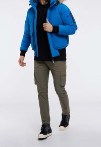 Man som bär en blå vinterjacka med huva, svart skjorta, olivgröna cargobyxor och svarta högskaftade sneakers som går på en vit bakgrund.