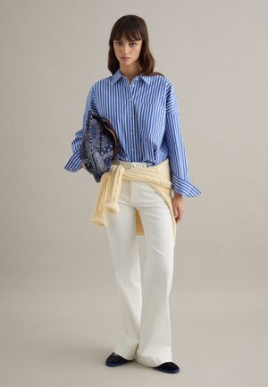 Mujer con camisa a rayas azules y blancas, suéter crema atado a la cintura, pantalones blancos, zapatos negros, sosteniendo un bolso de mano azul marino con estampado.